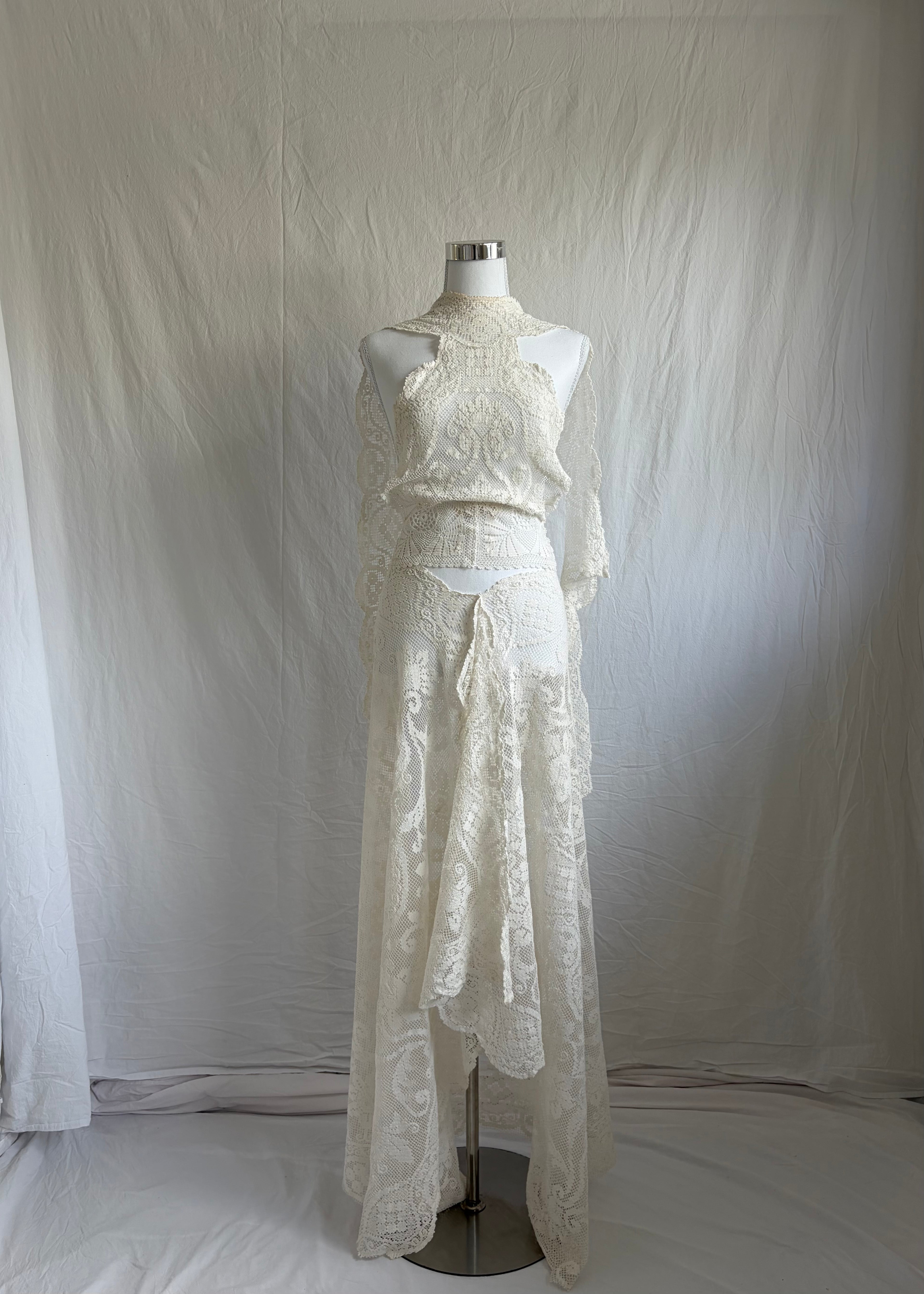 Ivory or cream lace two-piece set with long sleeves. The top features a high mock neck, an open shoulder/keyhole cutout design, and flared lace sleeves. The skirt is a matching high-waisted, asymmetrical maxi length with a handkerchief hem.