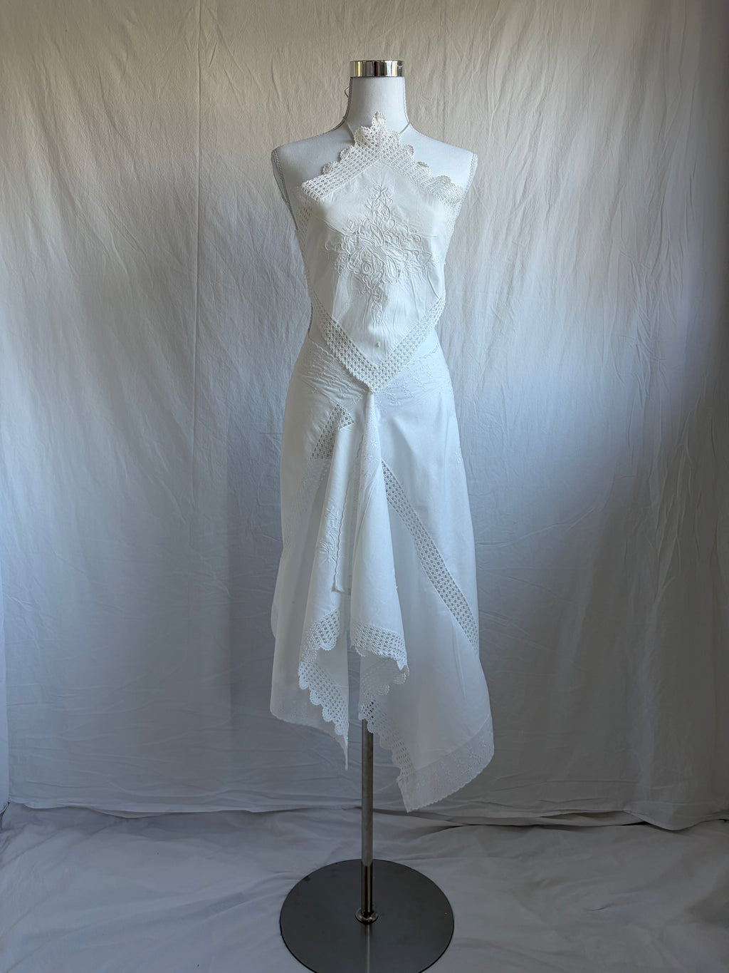 White upcycled two-piece set featuring vintage embroidered fabric. The set includes a diamond-shaped halter top with lace trim and an asymmetrical, midi-length wrap skirt with scalloped crochet lace edges and a crisscross waist tie.