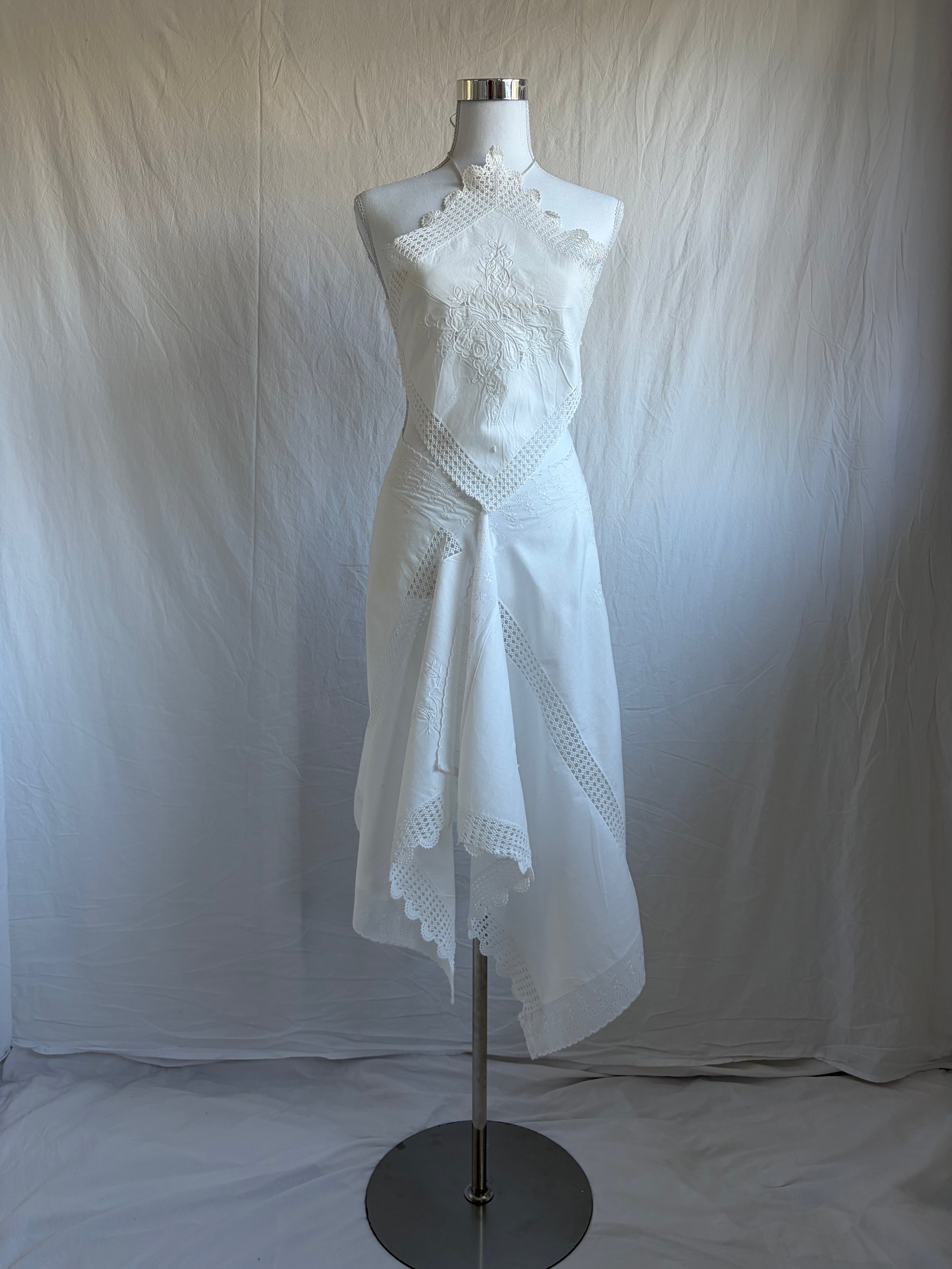 White upcycled two-piece set featuring vintage embroidered fabric. The set includes a diamond-shaped halter top with lace trim and an asymmetrical, midi-length wrap skirt with scalloped crochet lace edges and a crisscross waist tie.