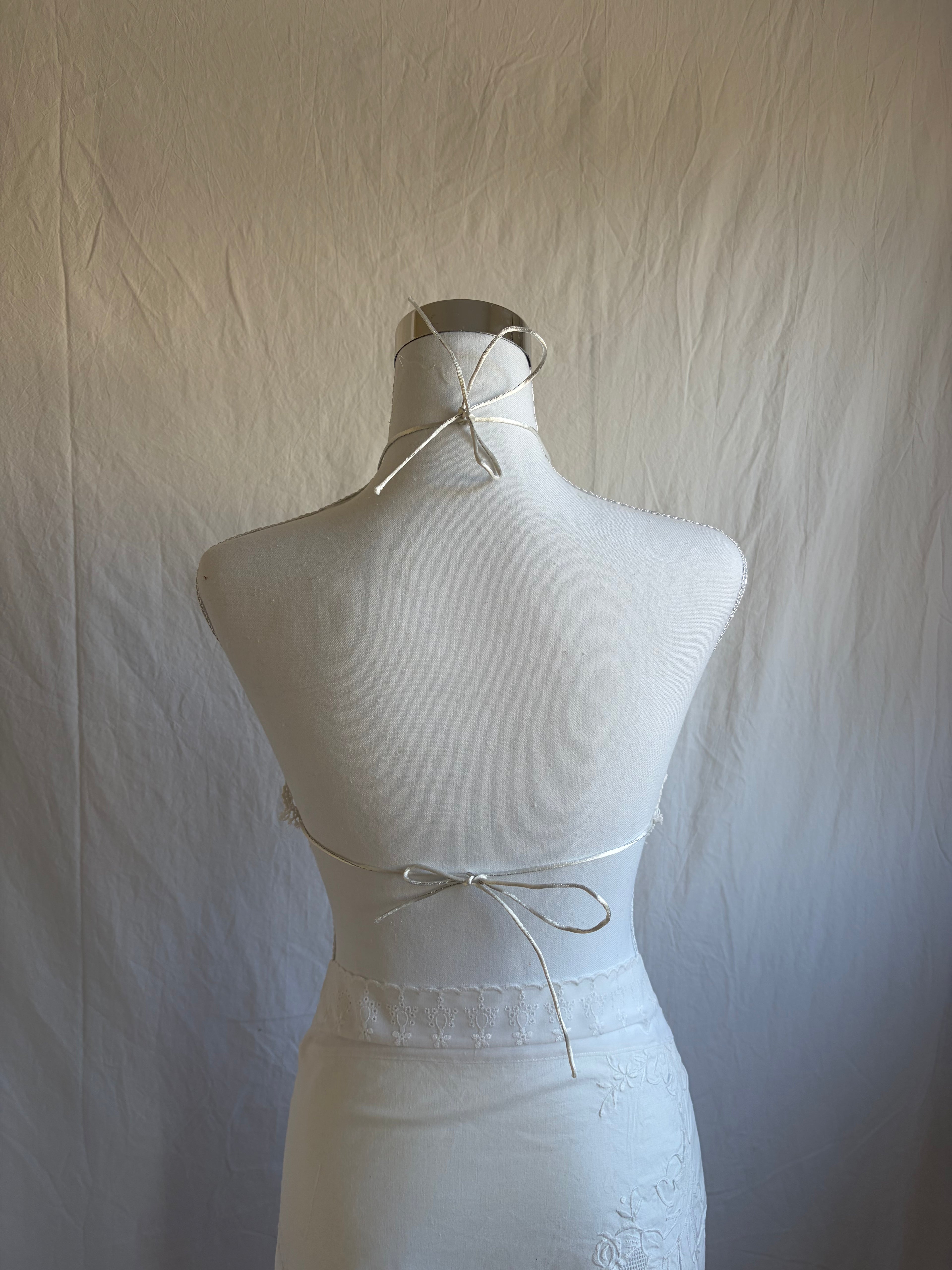 White upcycled two-piece set featuring vintage embroidered fabric. The set includes a diamond-shaped halter top with lace trim and an asymmetrical, midi-length wrap skirt with scalloped crochet lace edges and a crisscross waist tie.