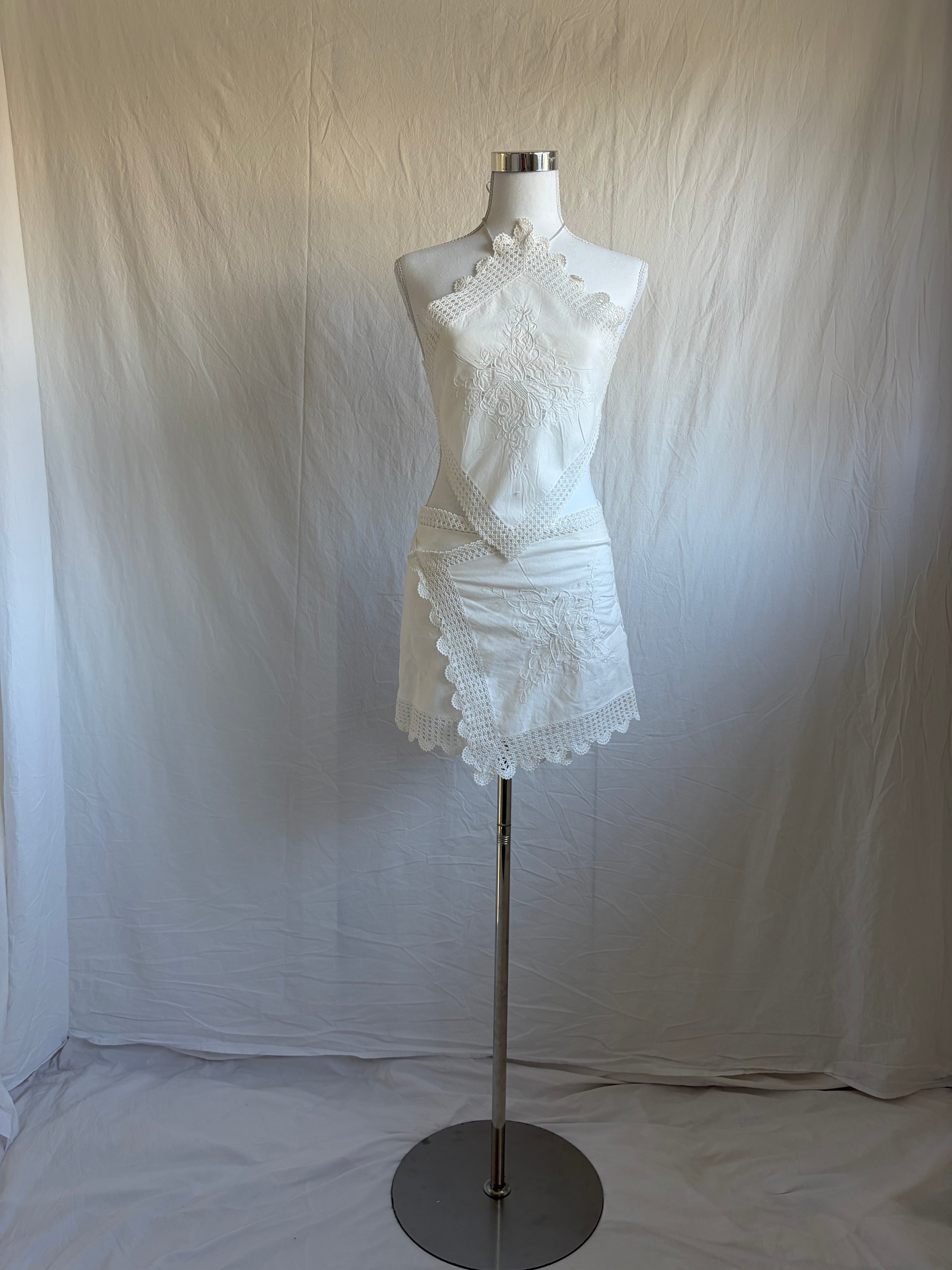 White upcycled two-piece set featuring vintage embroidered handkerchiefs. The set includes a diamond-shaped halter-neck top with scalloped crochet lace edges and a mini wrap skirt with a matching scalloped lace hem.