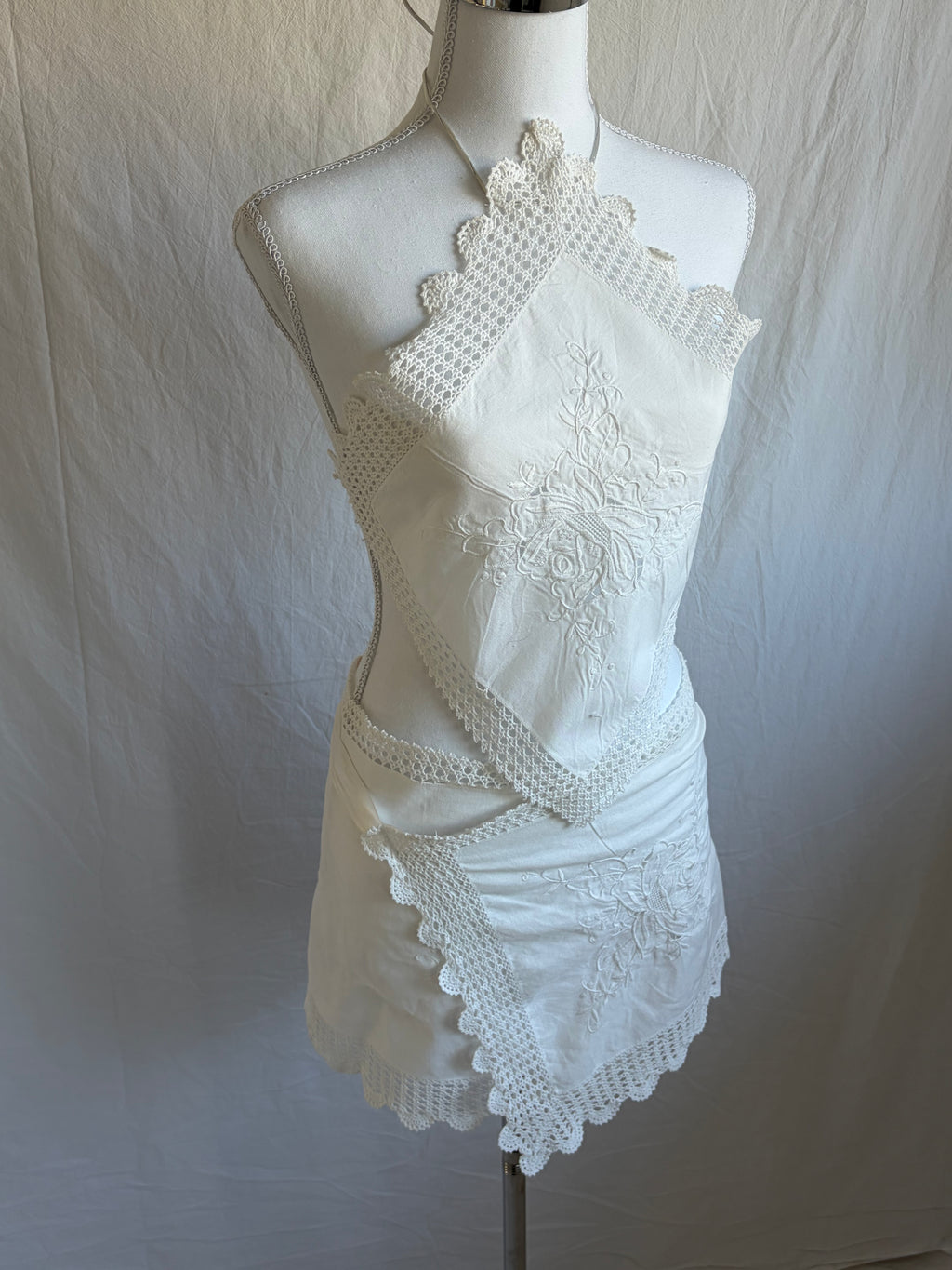 White upcycled two-piece set featuring vintage embroidered handkerchiefs. The set includes a diamond-shaped halter-neck top with scalloped crochet lace edges and a mini wrap skirt with a matching scalloped lace hem.