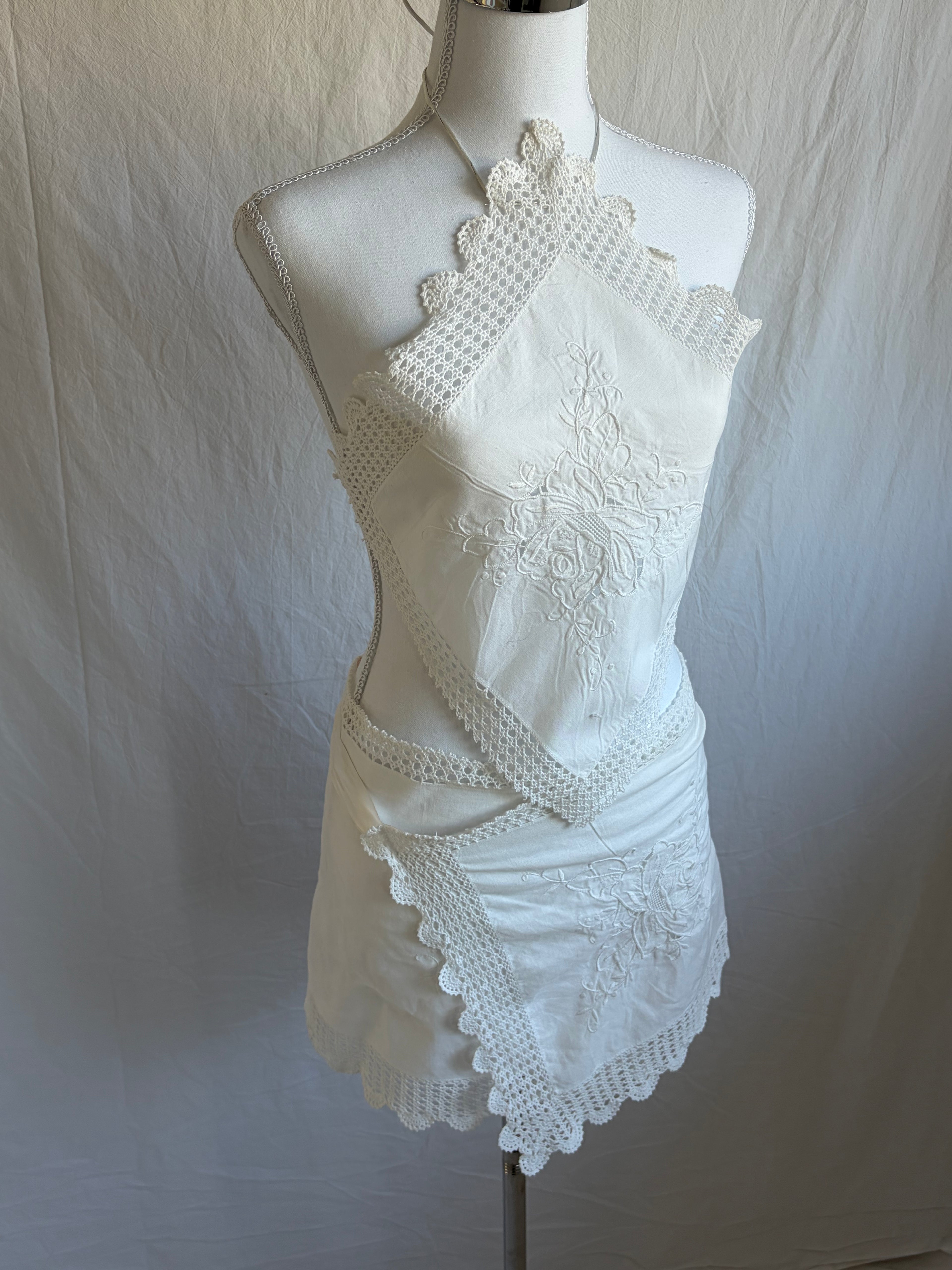 White upcycled two-piece set featuring vintage embroidered handkerchiefs. The set includes a diamond-shaped halter-neck top with scalloped crochet lace edges and a mini wrap skirt with a matching scalloped lace hem.
