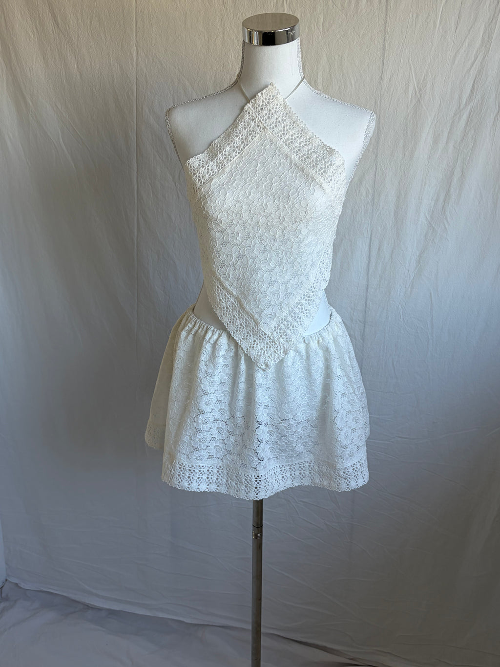 White two-piece set featuring a diamond-shaped halter-neck cropped top and a short, gathered mini skirt. Both pieces are made from a white floral lace fabric with a subtle eyelet pattern.