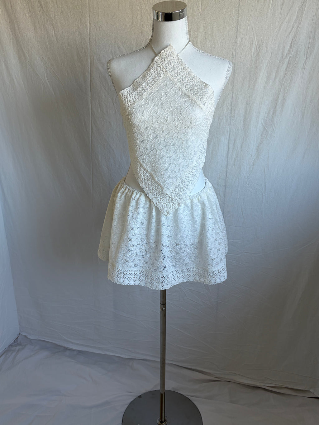 White two-piece set featuring a diamond-shaped halter-neck cropped top and a short, gathered mini skirt. Both pieces are made from a white floral lace fabric with a subtle eyelet pattern.