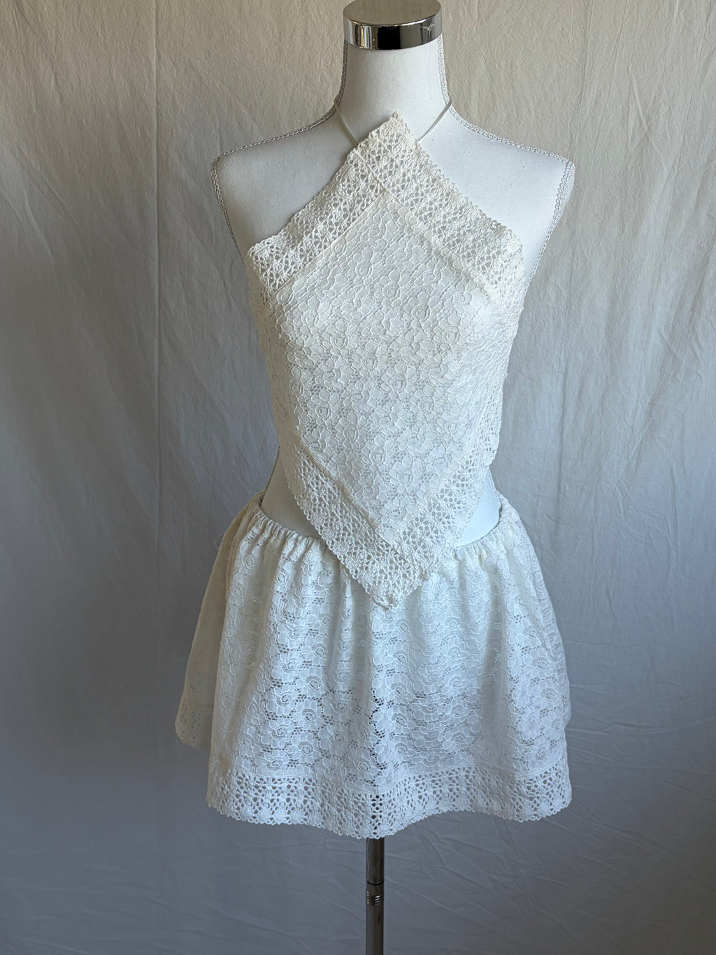 White two-piece set featuring a diamond-shaped halter-neck cropped top and a short, gathered mini skirt. Both pieces are made from a white floral lace fabric with a subtle eyelet pattern.