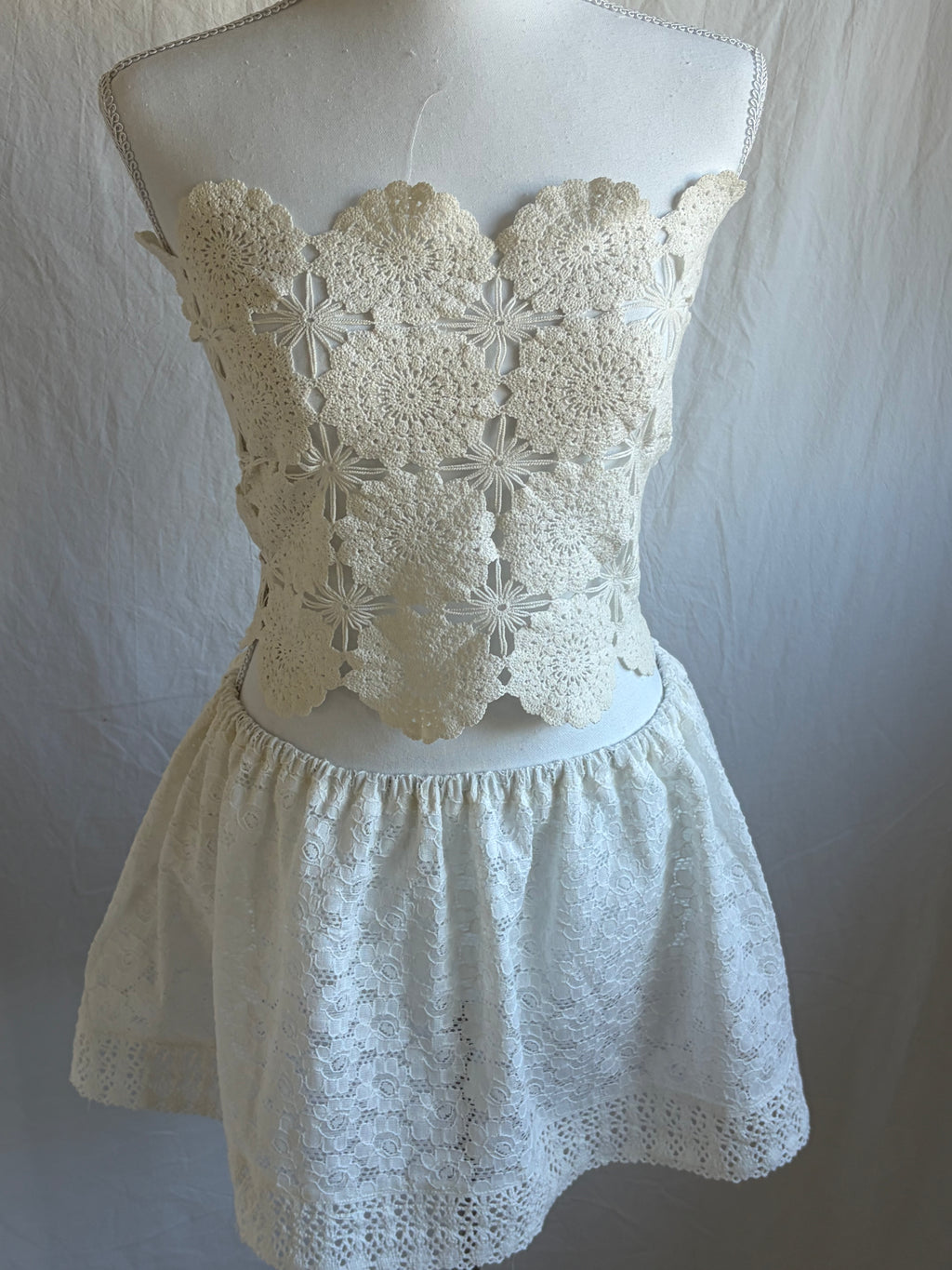 Text: Cream white two-piece set featuring a cropped, scalloped-edge, crochet floral lace bustier top and a high-waisted, short, elastic-waist skirt with eyelet lace fabric.
