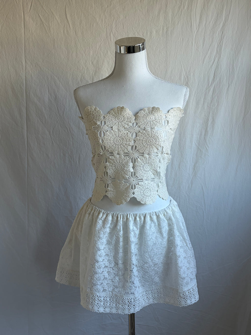 Cream white two-piece set featuring a cropped, scalloped-edge, crochet floral lace bustier top and a high-waisted, short, elastic-waist skirt with eyelet lace fabric.