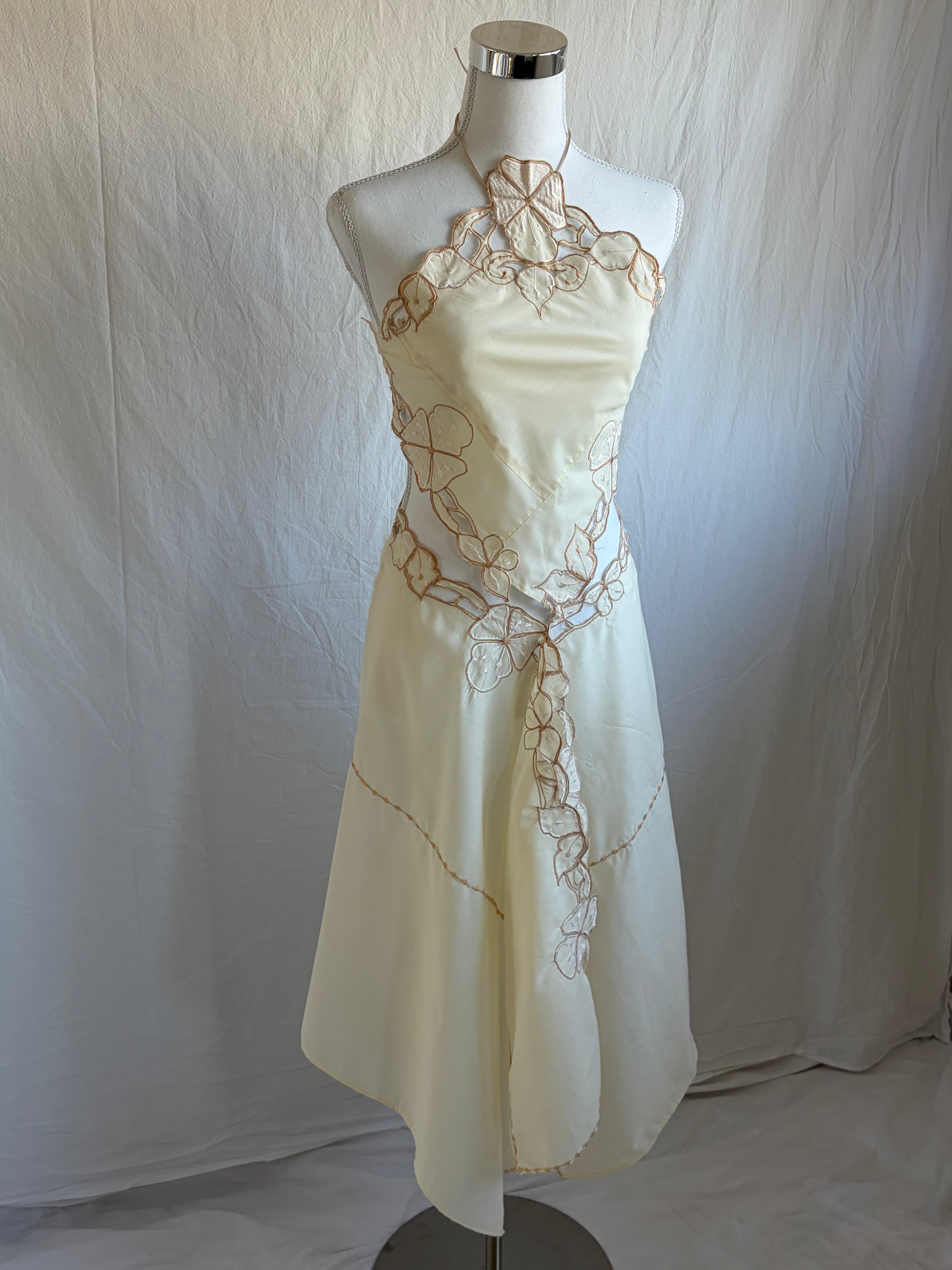 Cream or light yellow two-piece halter dress set with visible stitching and embroidery. The set features a handkerchief-style cropped halter top and an asymmetrical, midi-length skirt, both accented with a tan or gold floral embroidery tracing the seams.