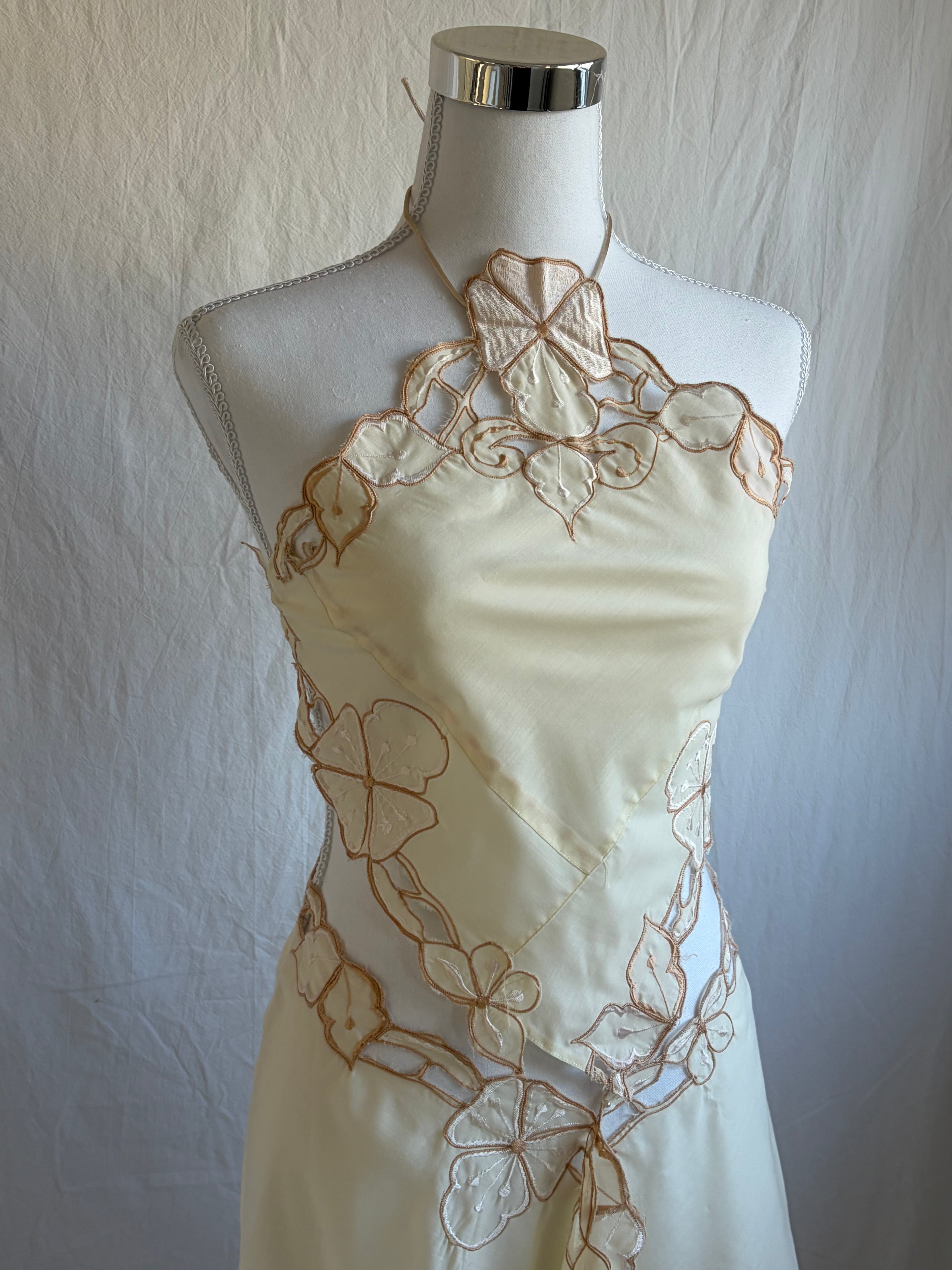Cream or light yellow two-piece halter dress set with visible stitching and embroidery. The set features a handkerchief-style cropped halter top and an asymmetrical, midi-length skirt, both accented with a tan or gold floral embroidery tracing the seams.
