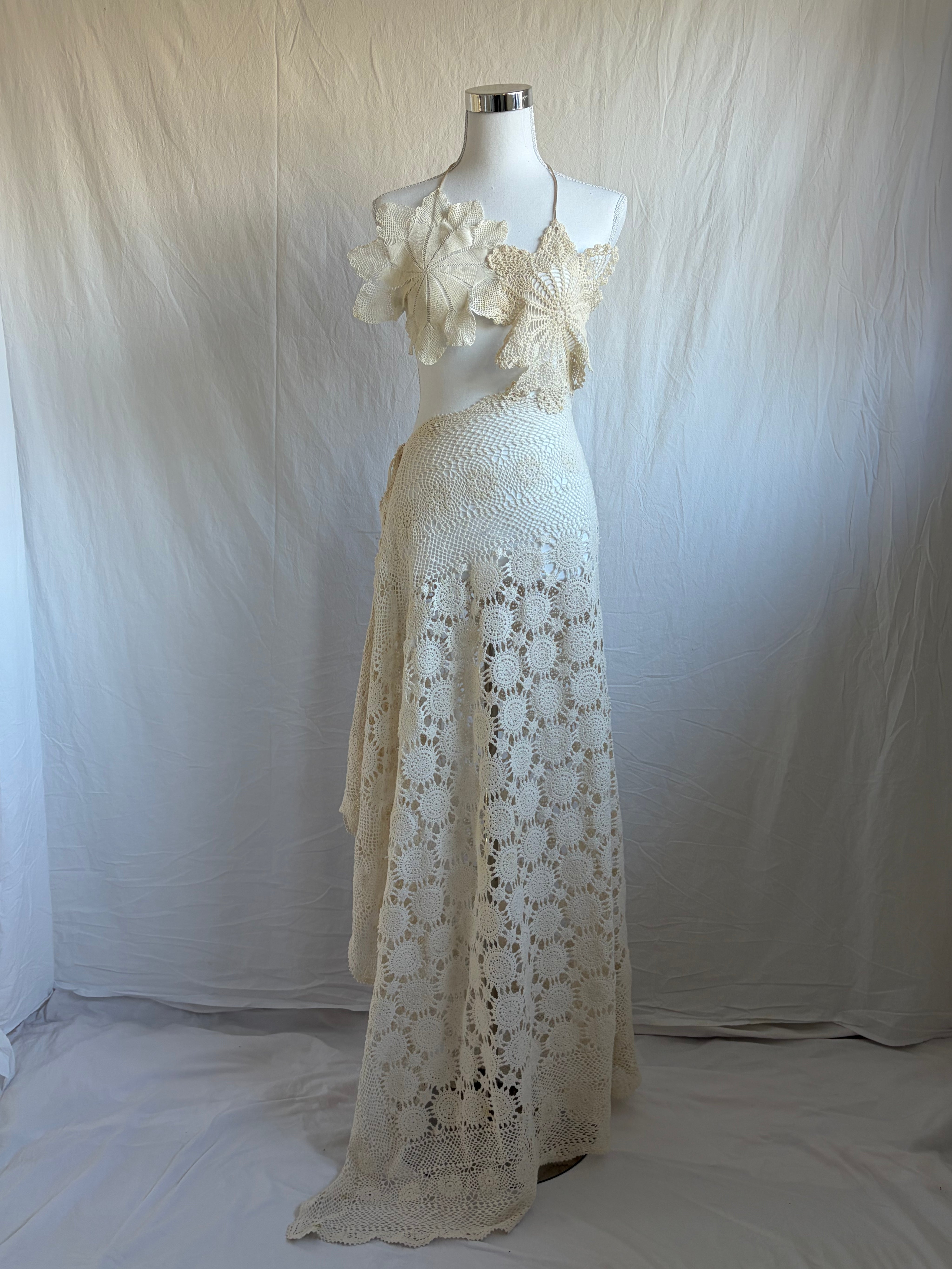 Vintage-style cream crochet two-piece set. The set includes a cropped halter top adorned with oversized three-dimensional floral appliqué details, and a high-waisted, asymmetrical maxi skirt featuring large circular crochet floral patterns.