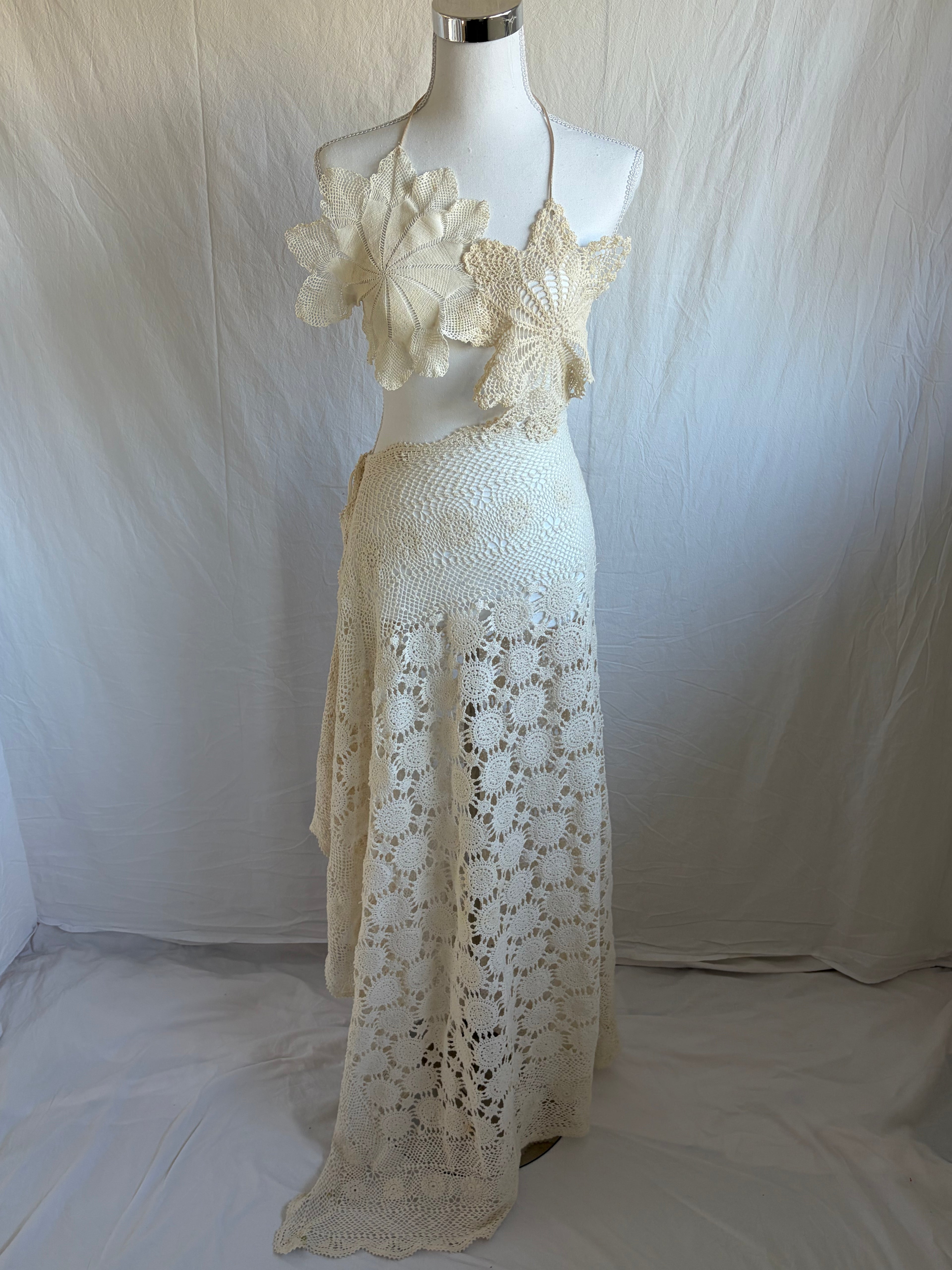 Vintage-style cream crochet two-piece set. The set includes a cropped halter top adorned with oversized three-dimensional floral appliqué details, and a high-waisted, asymmetrical maxi skirt featuring large circular crochet floral patterns.