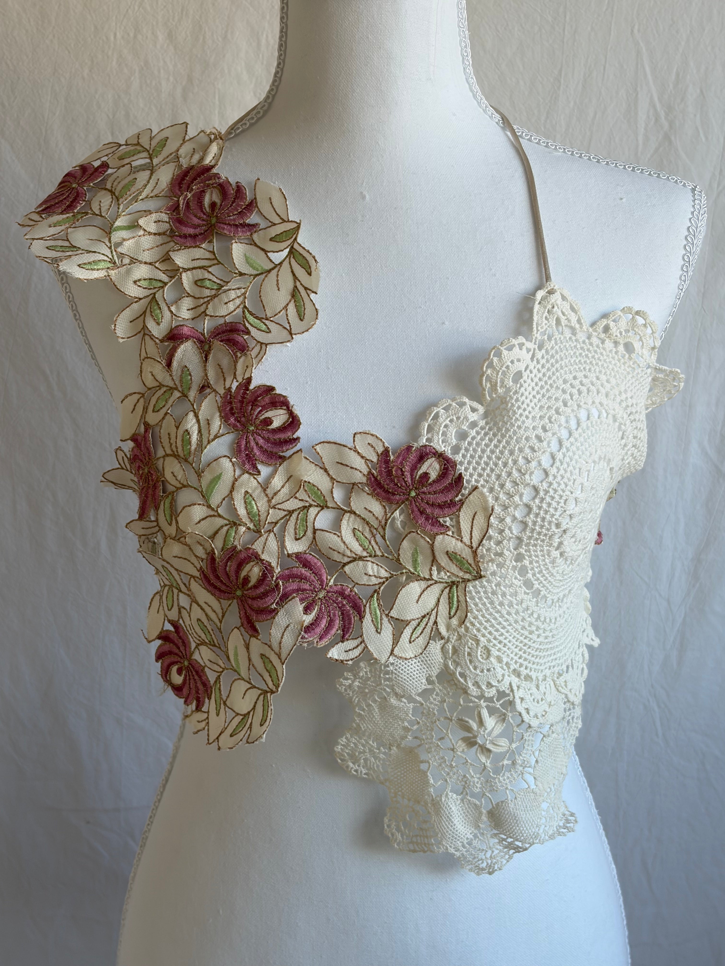 Model wearing a unique two-piece bohemian outfit. The top is a cropped halter made from cream crochet doilies and features an asymmetrical shoulder panel decorated with pink and cream floral fabric appliqués. It is paired with a sheer, flowing, high-waisted cream maxi skirt