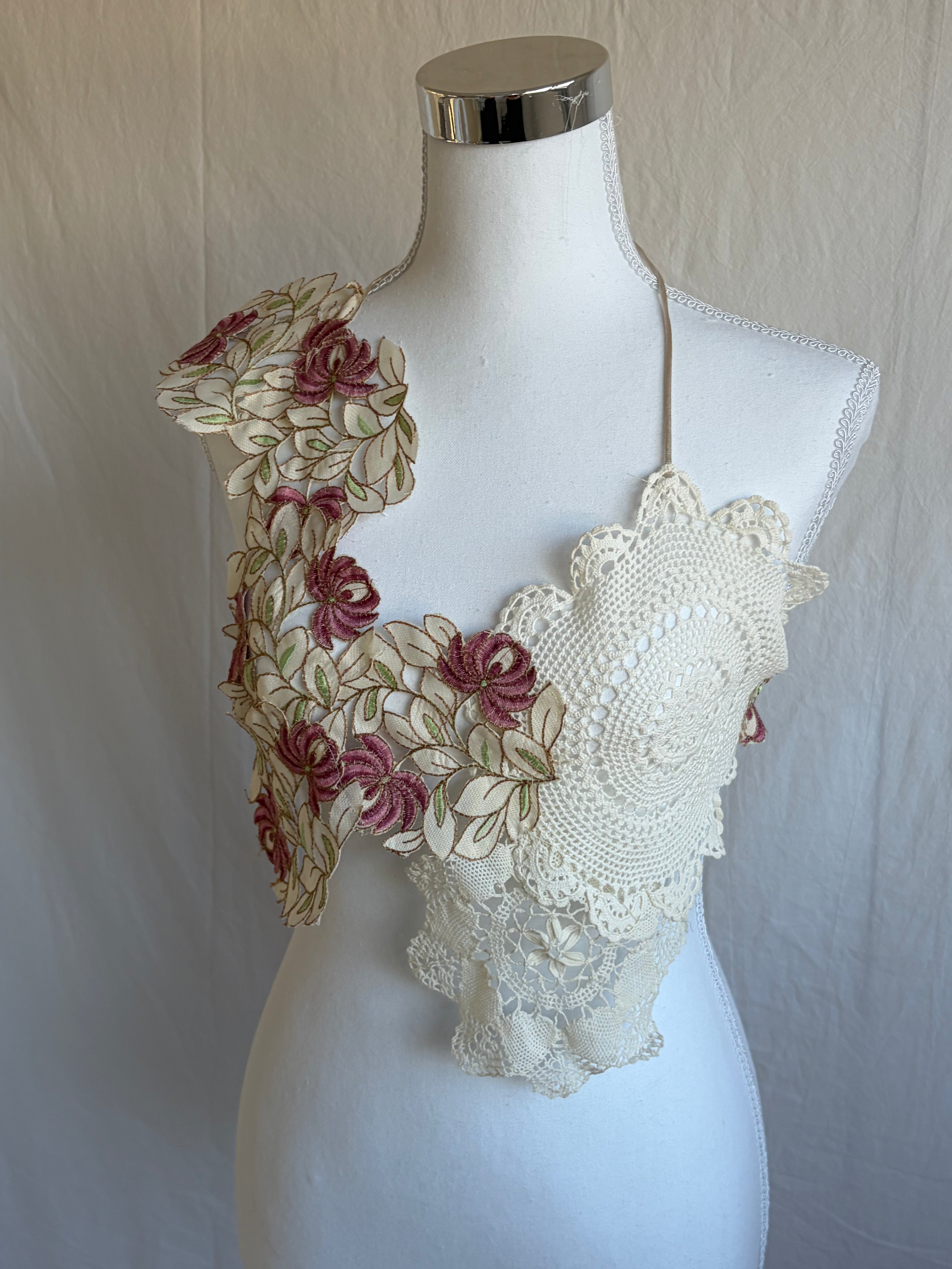 Model wearing a unique two-piece bohemian outfit. The top is a cropped halter made from cream crochet doilies and features an asymmetrical shoulder panel decorated with pink and cream floral fabric appliqués. It is paired with a sheer, flowing, high-waisted cream maxi skirt