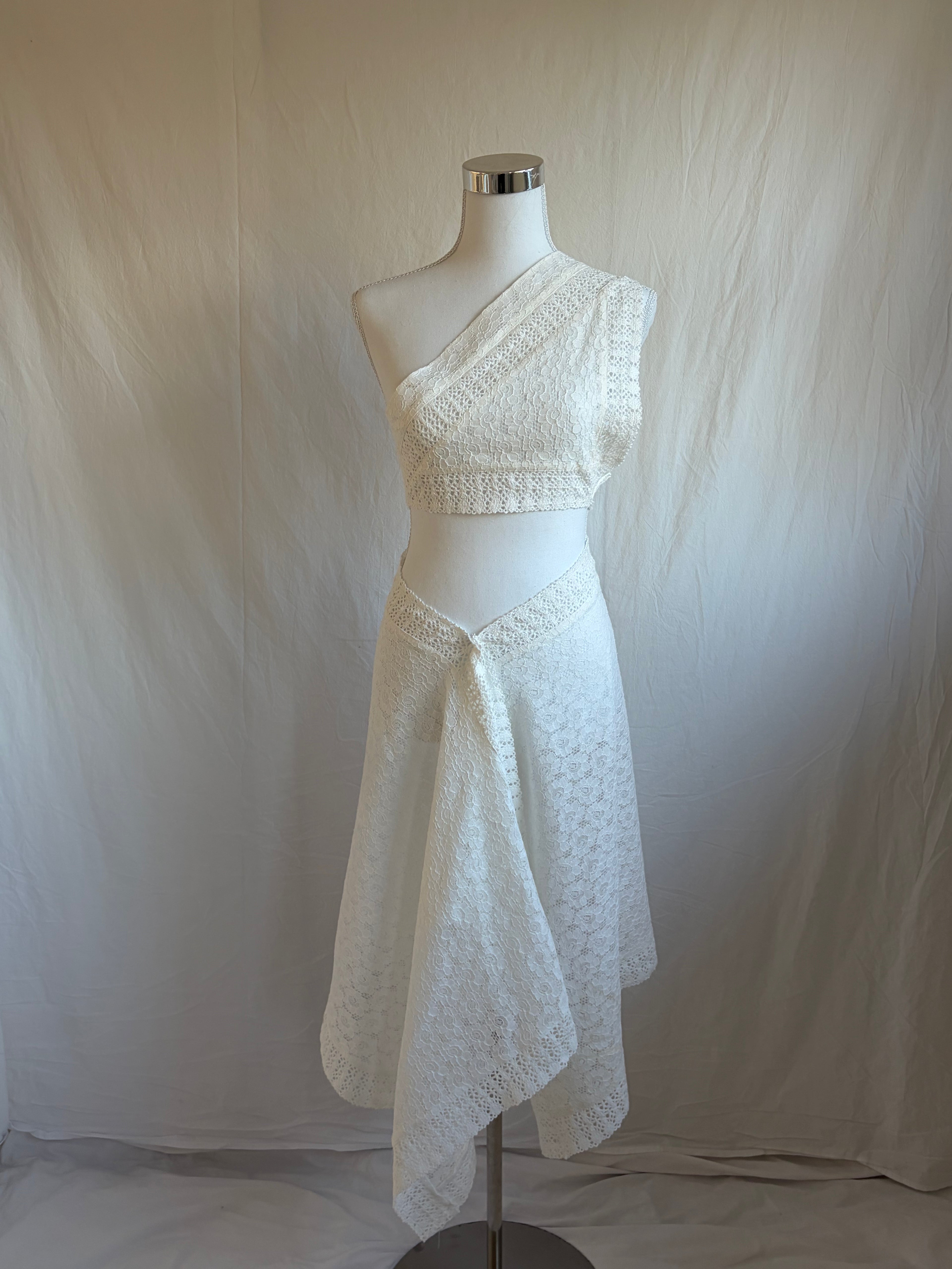 white two-piece set featuring an asymmetrical, one-shoulder cropped top and a midi-length handkerchief or draped wrap skirt. Both pieces are made from a white textured lace or eyelet fabric.