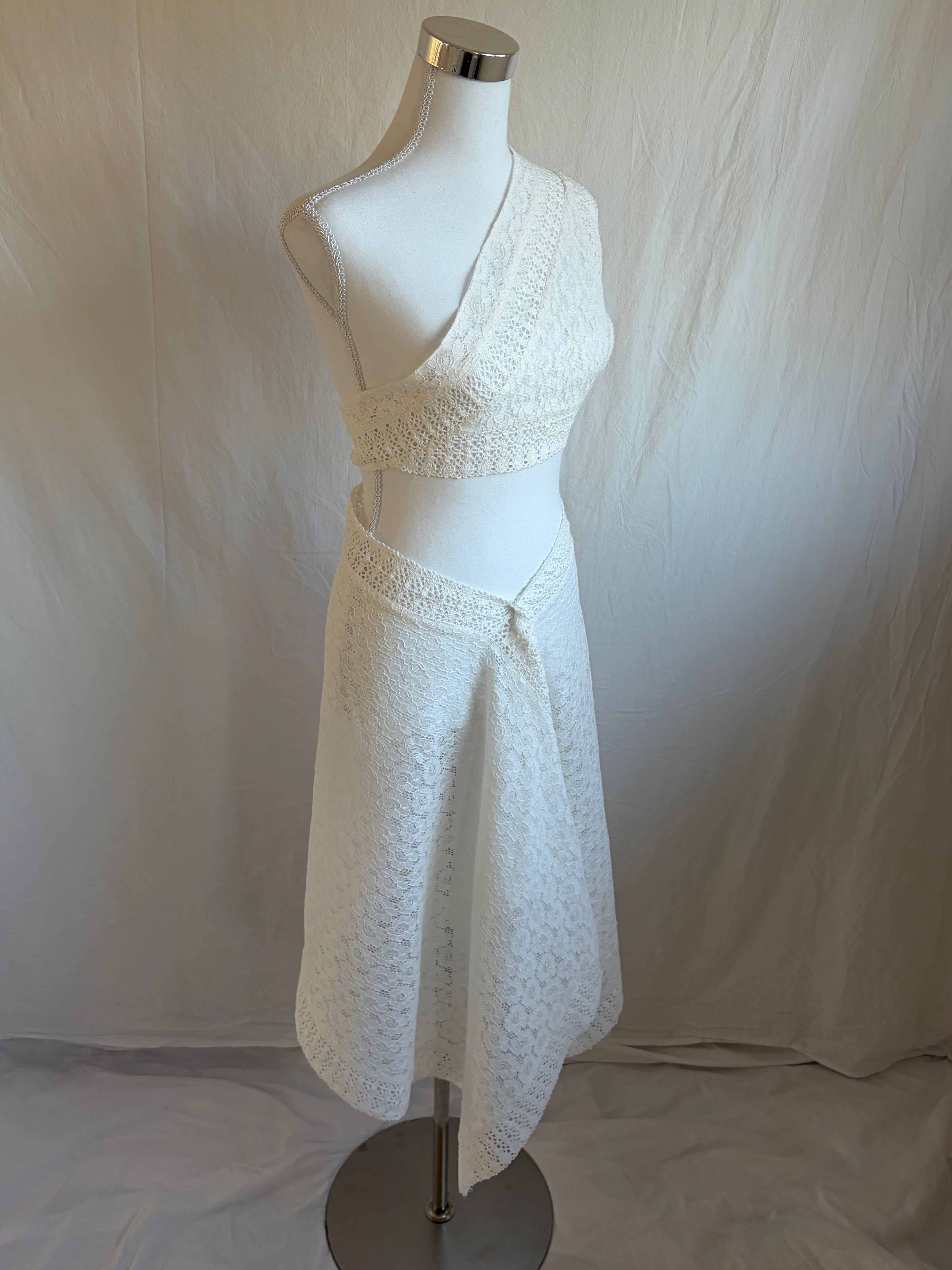 white two-piece set featuring an asymmetrical, one-shoulder cropped top and a midi-length handkerchief or draped wrap skirt. Both pieces are made from a white textured lace or eyelet fabric.