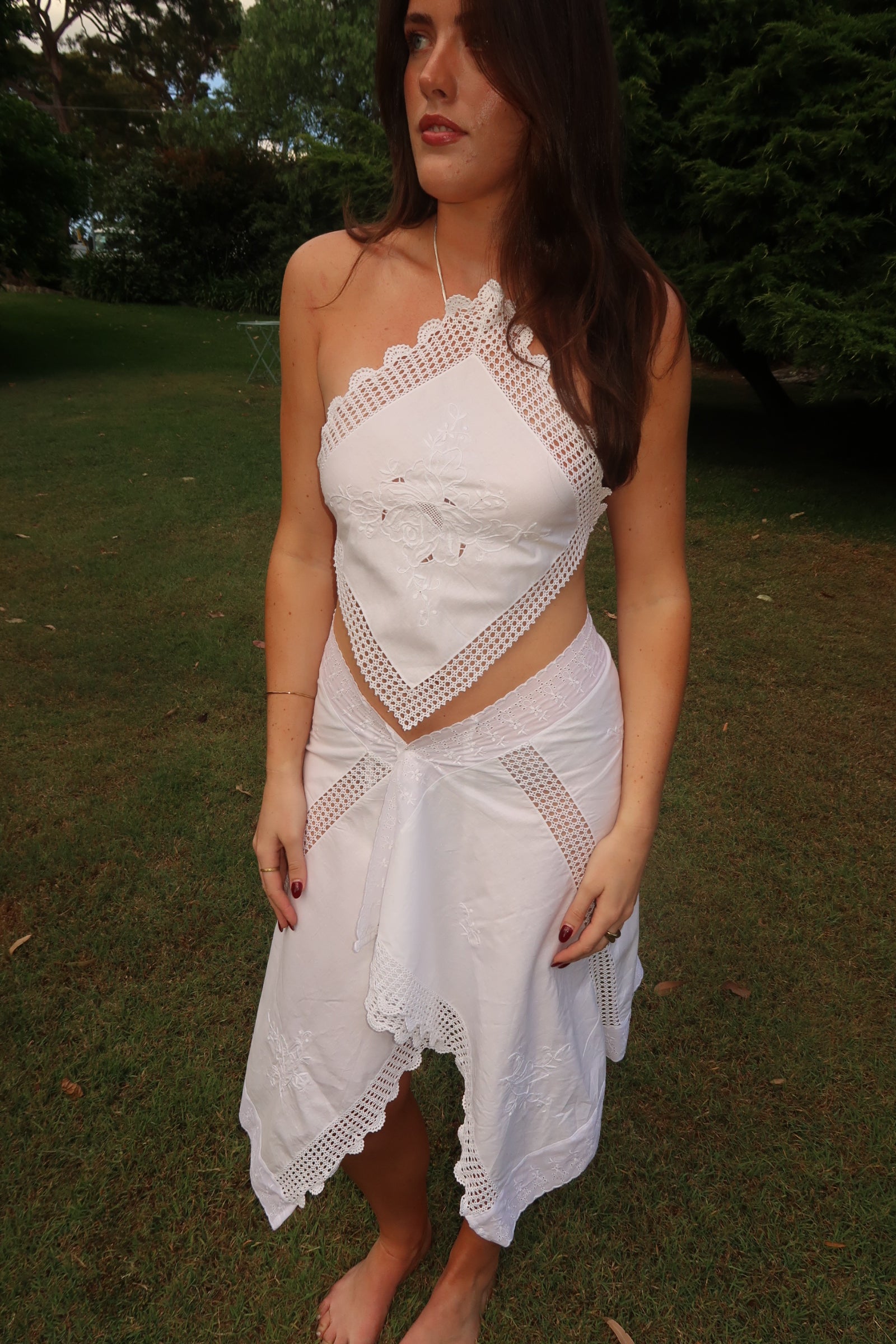 Model standing barefoot in a grassy outdoor setting wearing a white upcycled two-piece set. The top is an asymmetrical, one-shoulder halter style made from embroidered white fabric with a crocheted lace trim. The skirt is a matching asymmetrical midi-length wrap style with a lace-trimmed, scalloped hem.