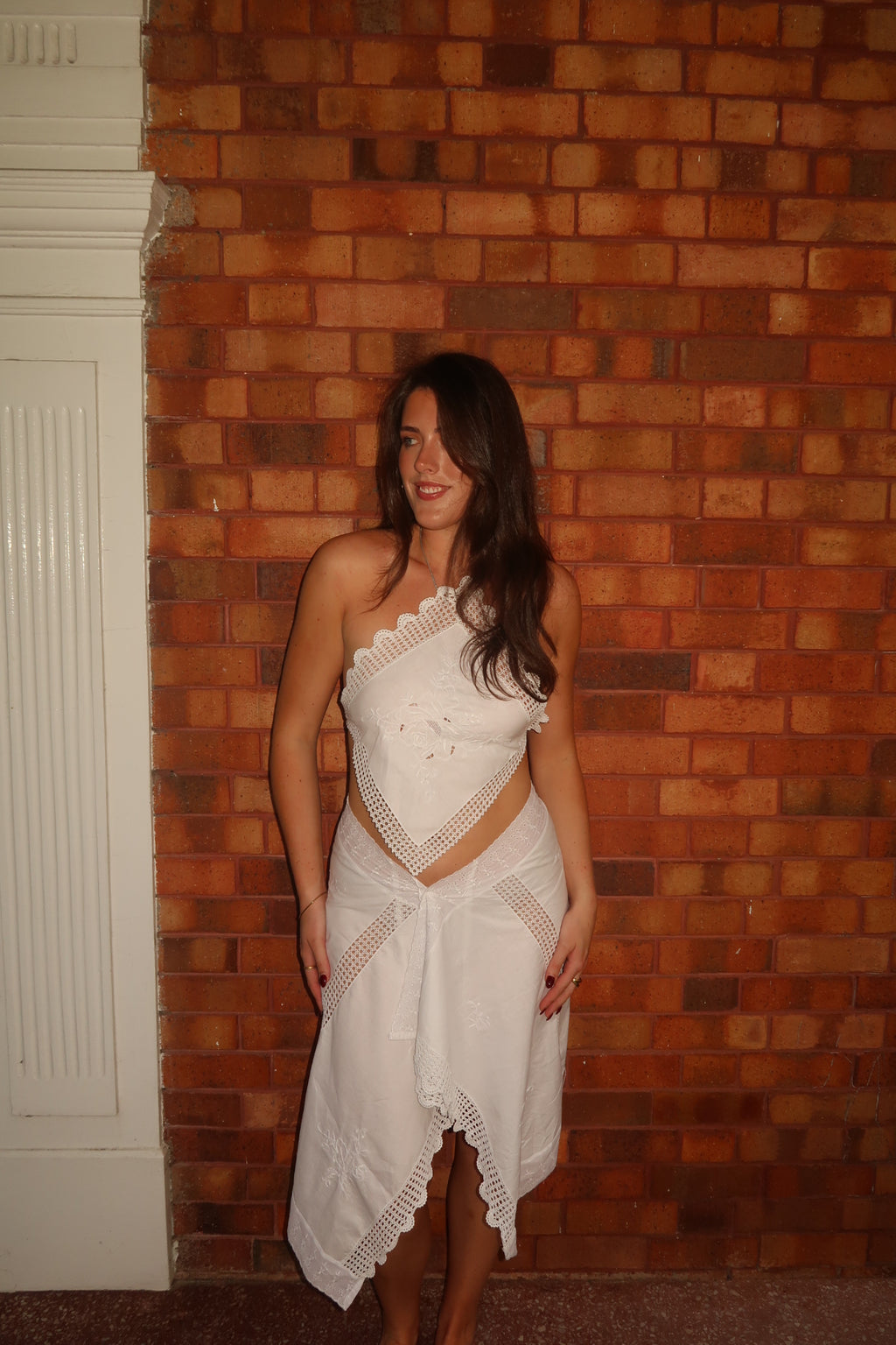 Model standing barefoot in a grassy outdoor setting wearing a white upcycled two-piece set. The top is an asymmetrical, one-shoulder halter style made from embroidered white fabric with a crocheted lace trim. The skirt is a matching asymmetrical midi-length wrap style with a lace-trimmed, scalloped hem.