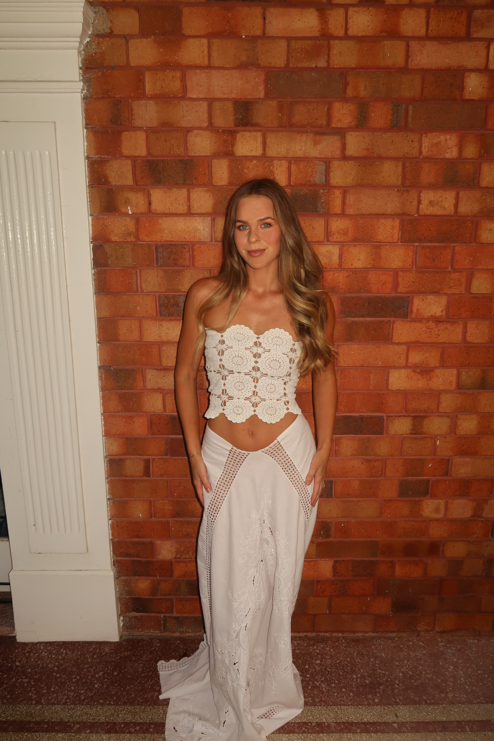 Model wearing a cream bohemian two-piece set against a red brick wall. The top is a cropped, scalloped-edge crochet bustier with floral medallion patterns. The bottom is a high-waisted, flowy maxi skirt with lace-up or cut-out details over the hips and subtle embroidery near the hem.