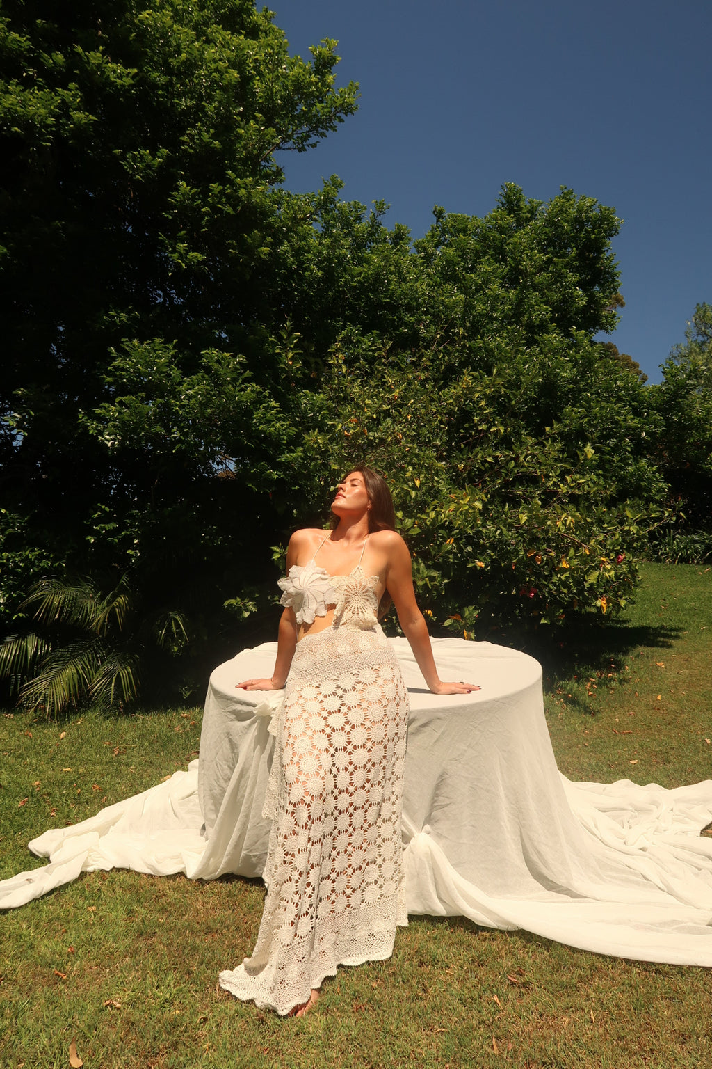 Vintage-style cream crochet two-piece set. The set includes a cropped halter top adorned with oversized three-dimensional floral appliqué details, and a high-waisted, asymmetrical maxi skirt featuring large circular crochet floral patterns.