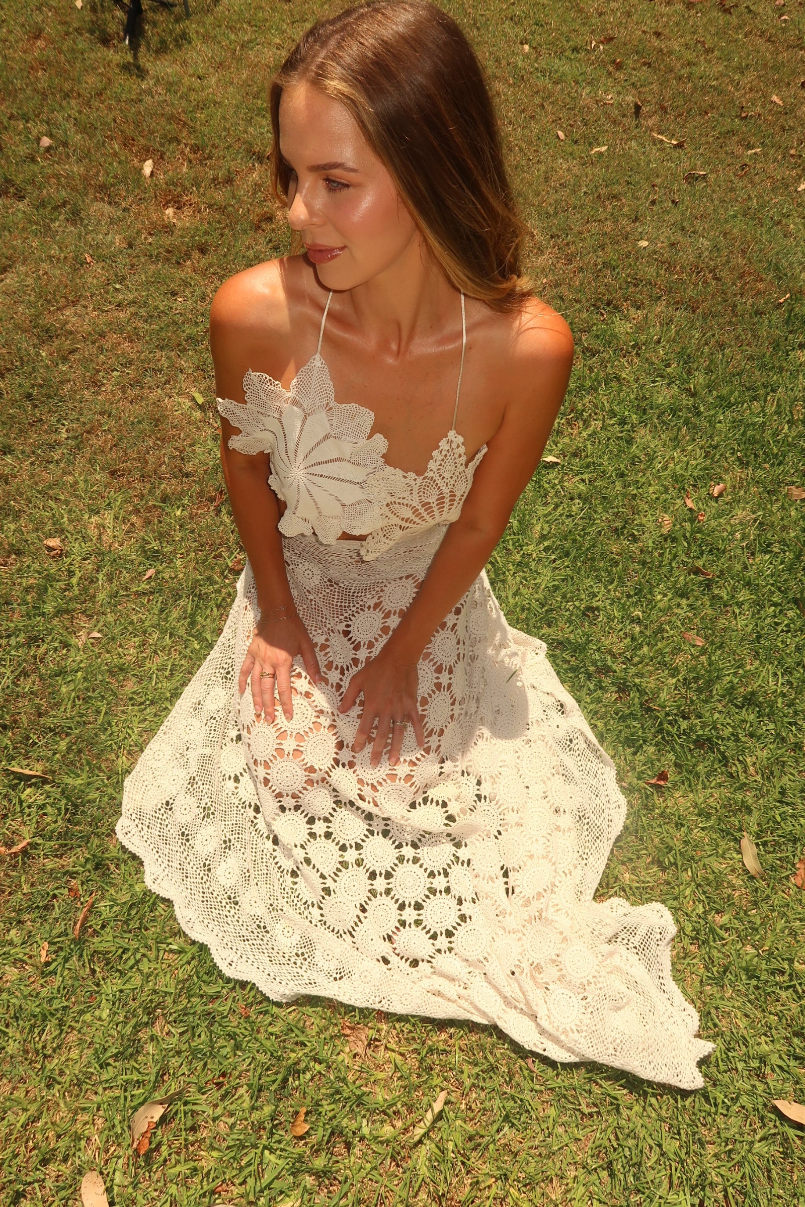 Vintage-style cream crochet two-piece set. The set includes a cropped halter top adorned with oversized three-dimensional floral appliqué details, and a high-waisted, asymmetrical maxi skirt featuring large circular crochet floral patterns.