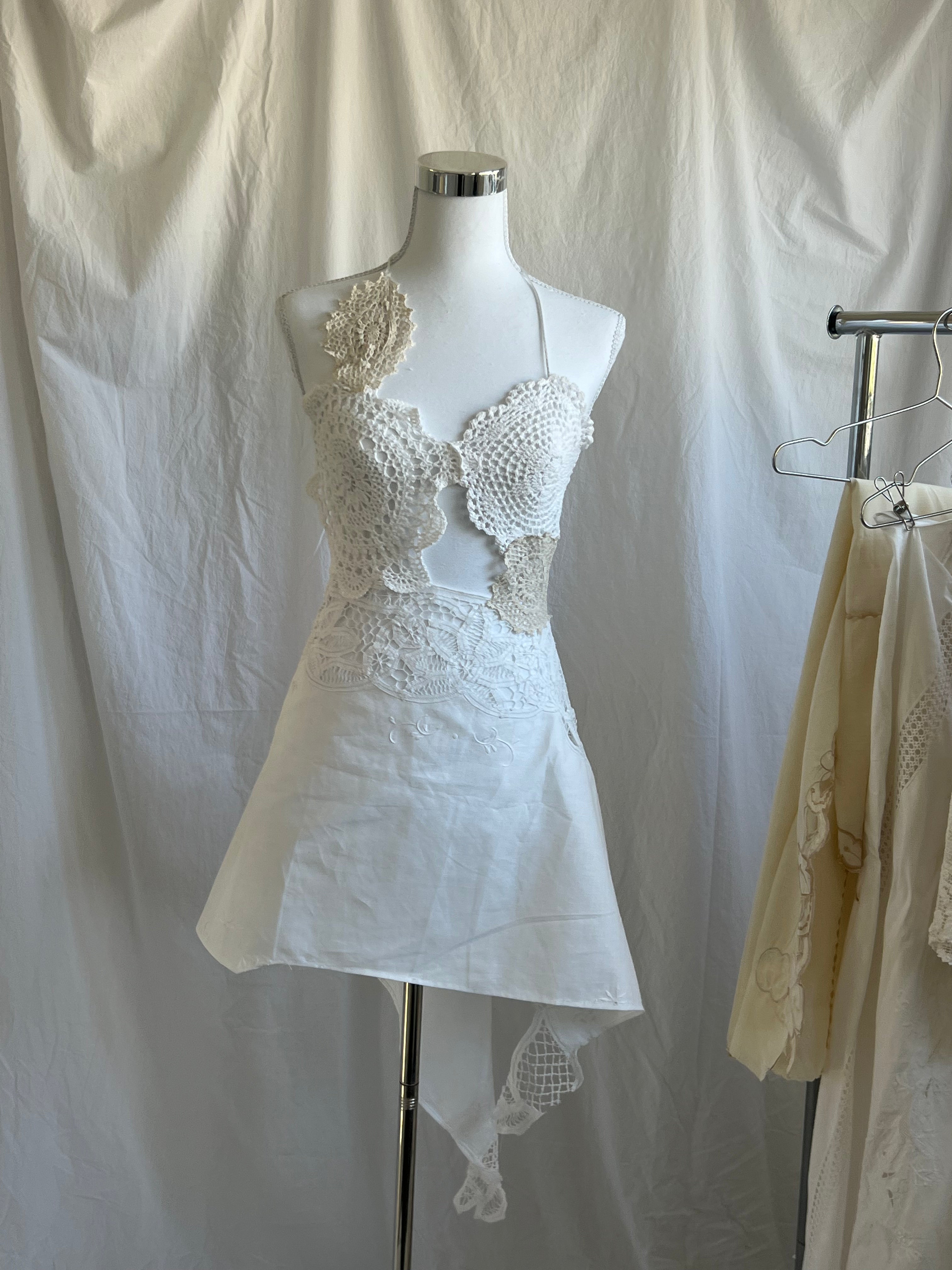 White two-piece set featuring a bustier-style crochet halter top with scallop details and a single floral appliqué on the shoulder. The skirt is a short, asymmetrical mini-skirt made of solid white fabric, with a contrasting white crochet lace waistline and a hanging crocheted fabric tail.