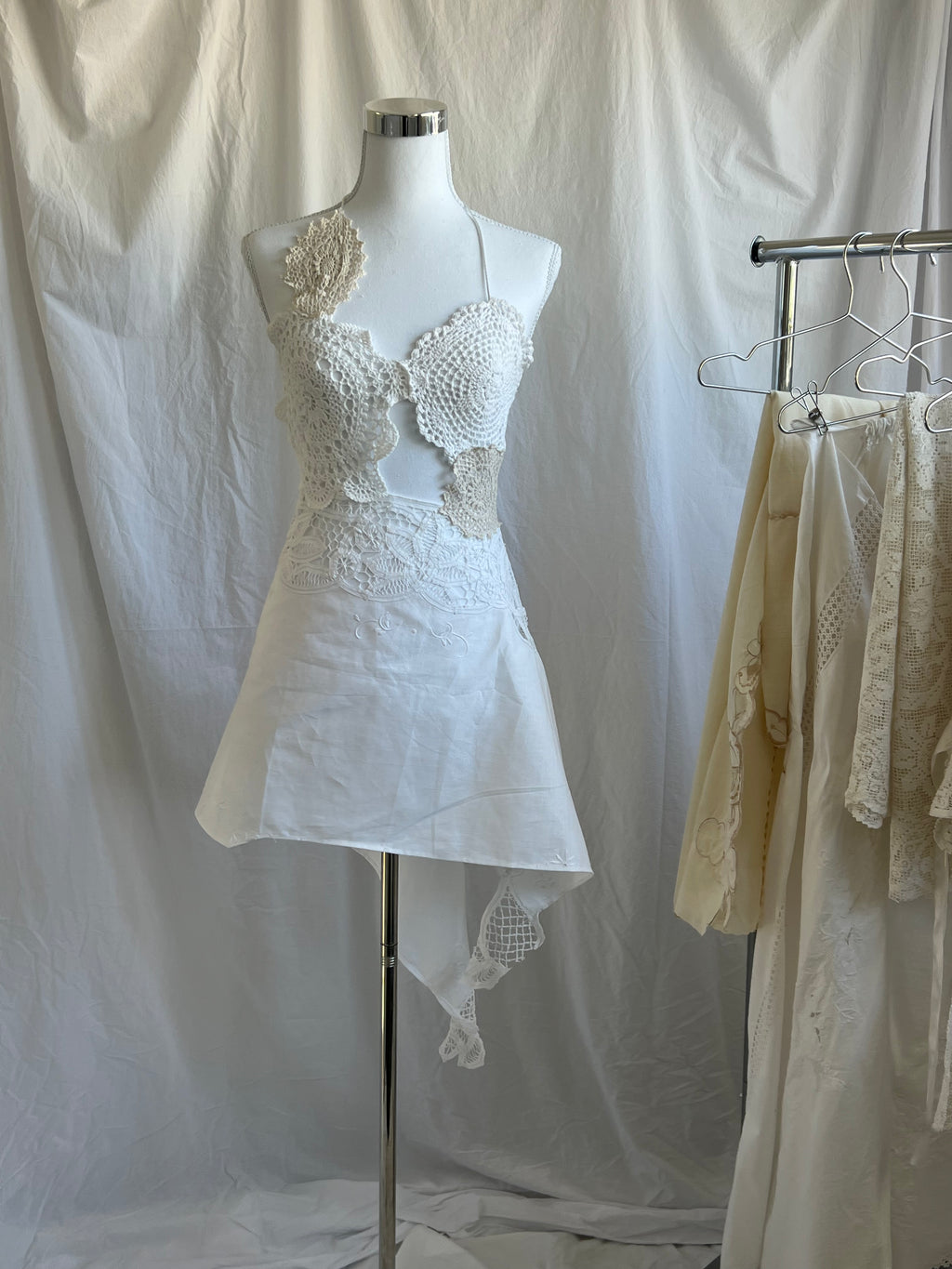 White two-piece set featuring a bustier-style crochet halter top with scallop details and a single floral appliqué on the shoulder. The skirt is a short, asymmetrical mini-skirt made of solid white fabric, with a contrasting white crochet lace waistline and a hanging crocheted fabric tail.