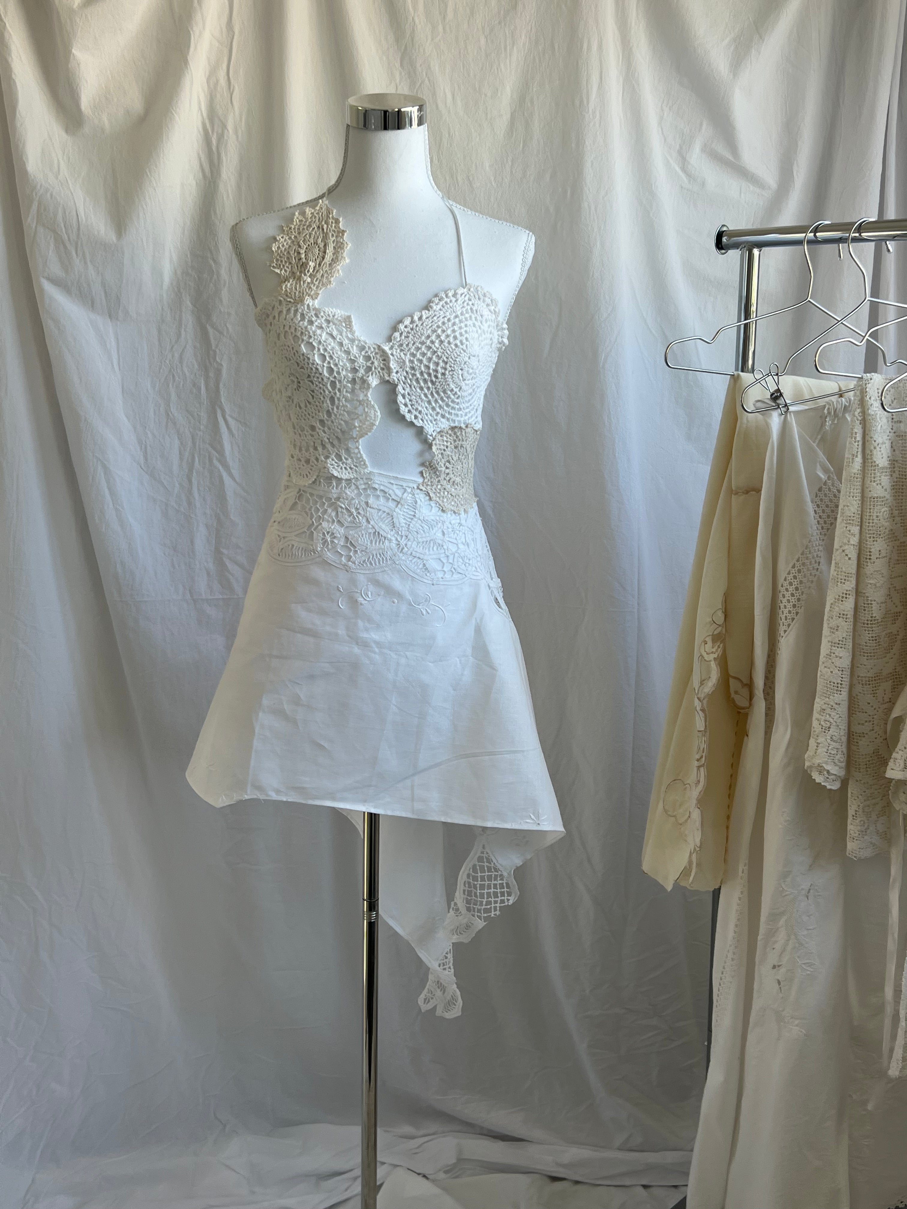 White two-piece set featuring a bustier-style crochet halter top with scallop details and a single floral appliqué on the shoulder. The skirt is a short, asymmetrical mini-skirt made of solid white fabric, with a contrasting white crochet lace waistline and a hanging crocheted fabric tail.