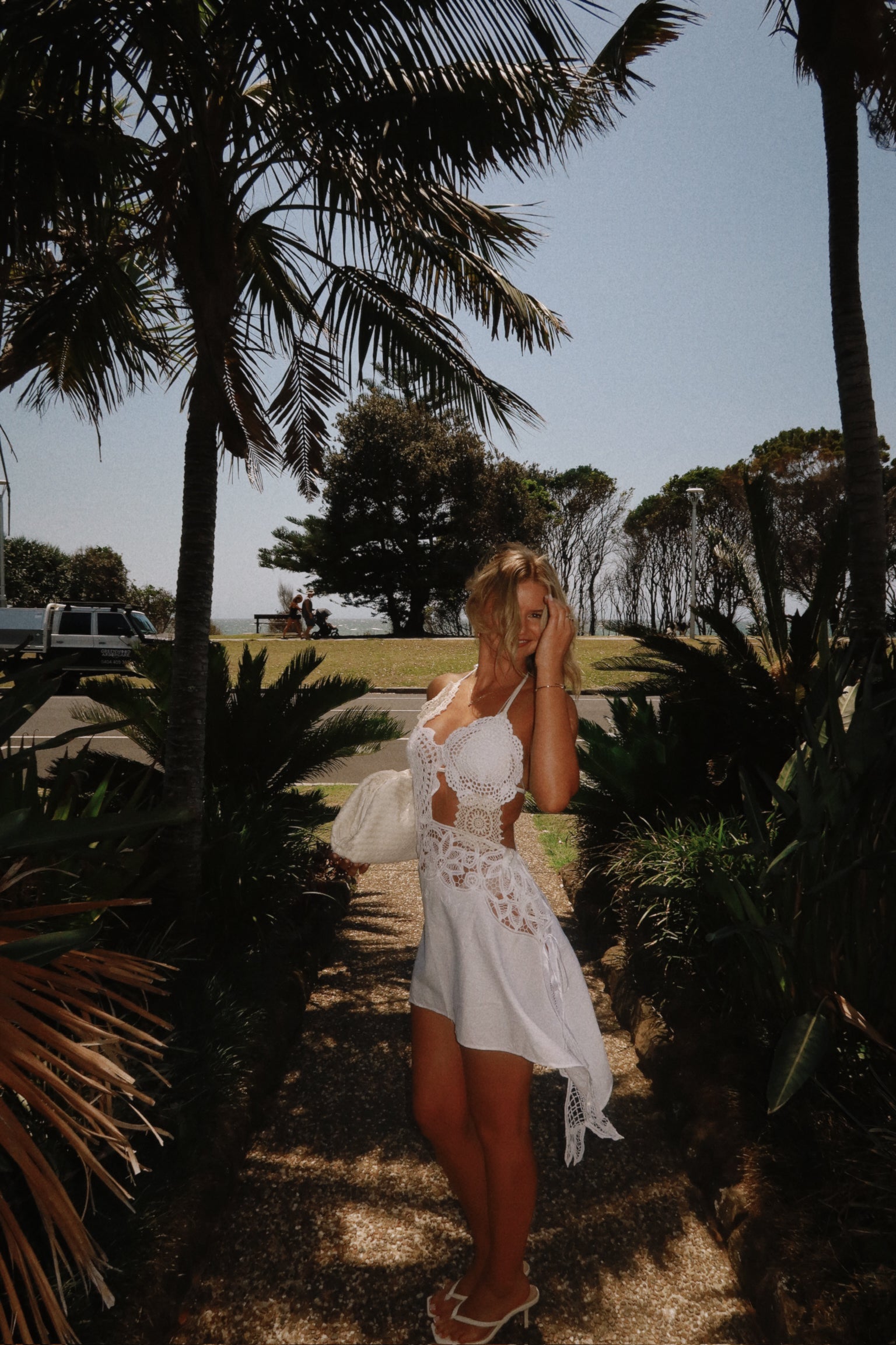 White two-piece set featuring a bustier-style crochet halter top with scallop details and a single floral appliqué on the shoulder. The skirt is a short, asymmetrical mini-skirt made of solid white fabric, with a contrasting white crochet lace waistline and a hanging crocheted fabric tail.
