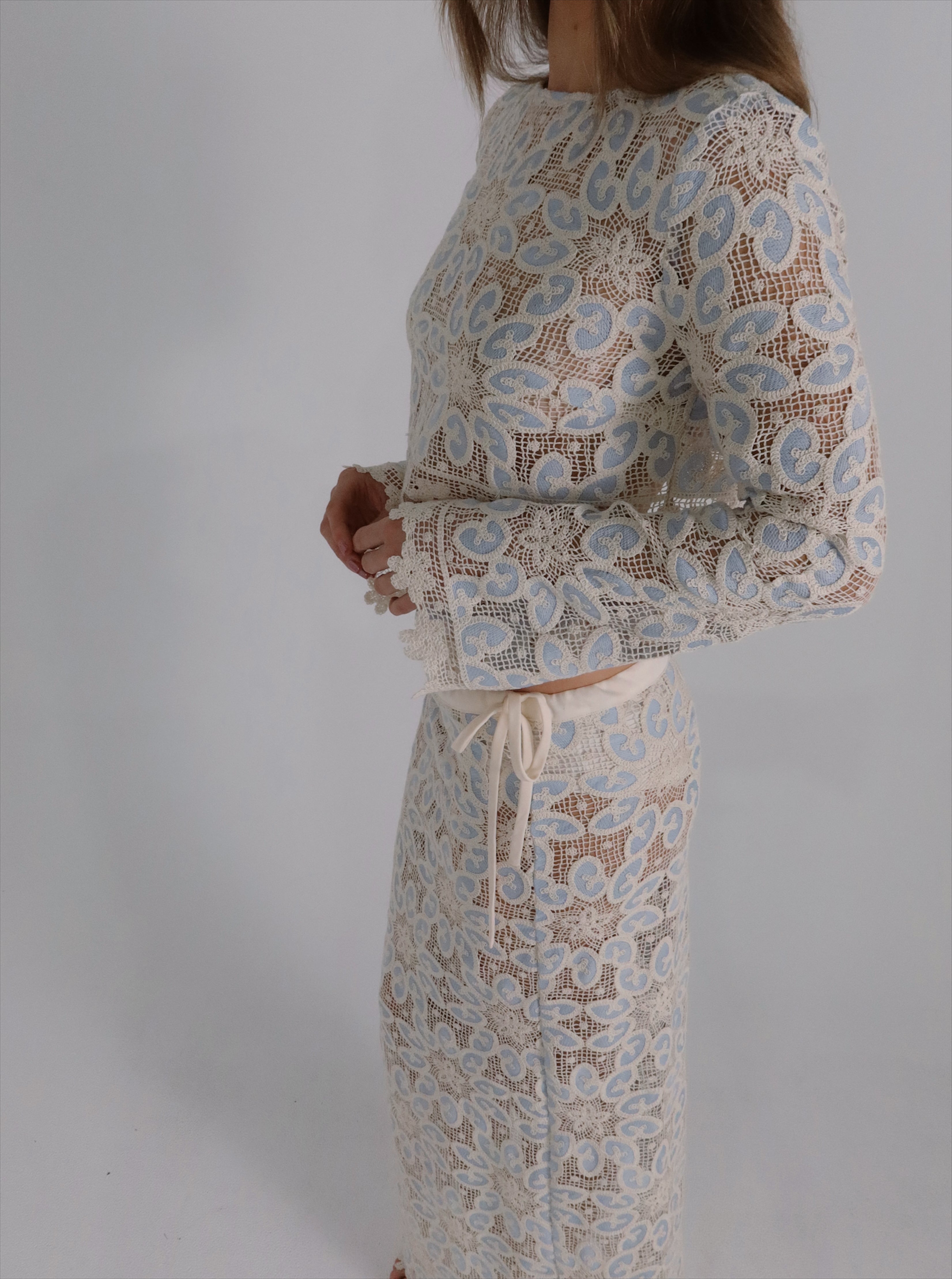 A model wearing a matching two-piece lace set consisting of a long-sleeve crop top and a high-waisted pencil skirt in a cream and light blue floral pattern.