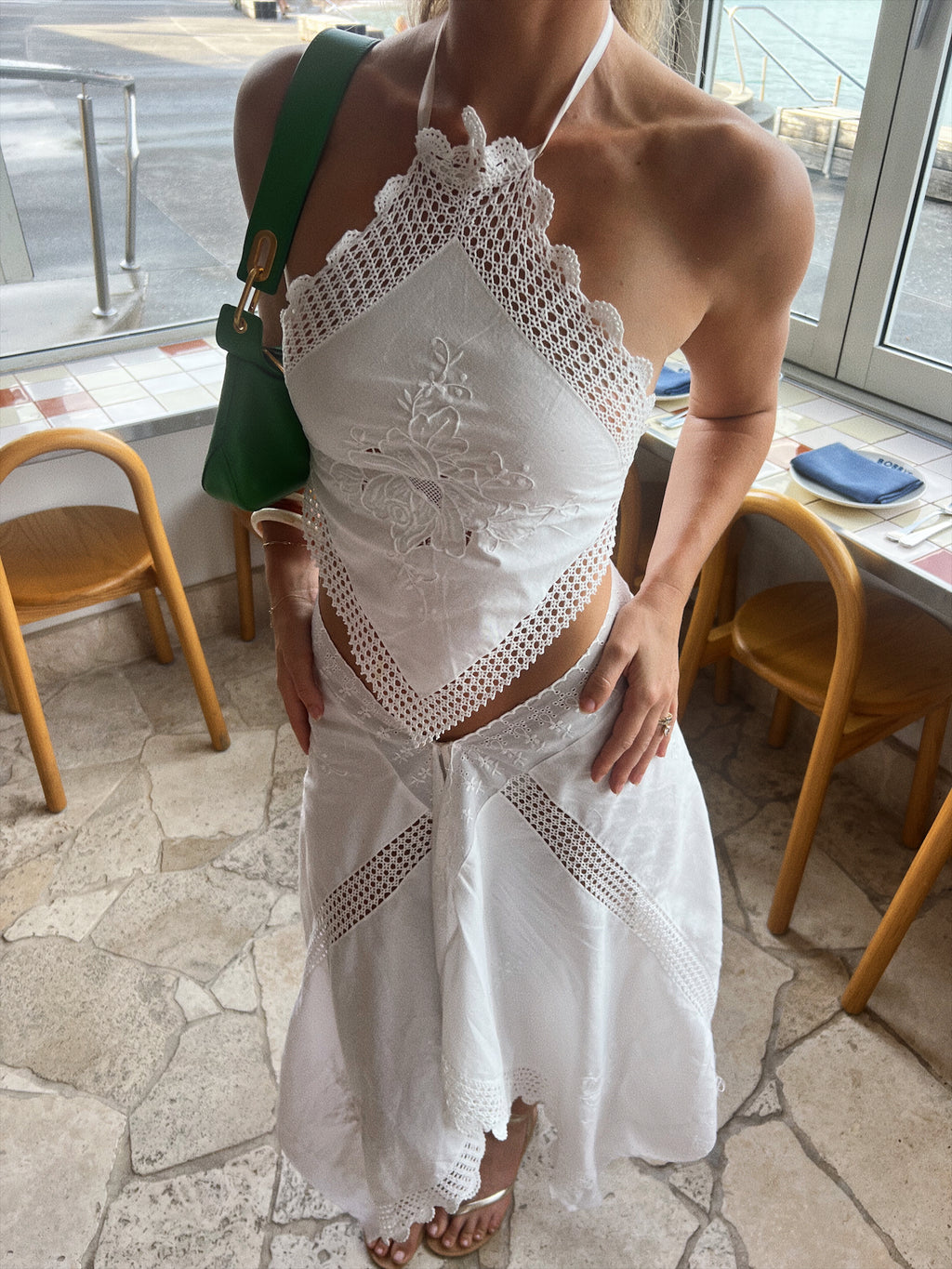 Model standing barefoot in a grassy outdoor setting wearing a white upcycled two-piece set. The top is an asymmetrical, one-shoulder halter style made from embroidered white fabric with a crocheted lace trim. The skirt is a matching asymmetrical midi-length wrap style with a lace-trimmed, scalloped hem.